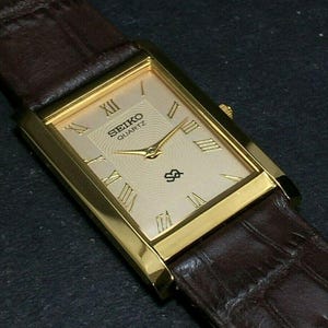 May include: A gold-toned Seiko Quartz watch with a rectangular face and Roman numeral hour markers. The watch has a brown leather band. The watch face is a light champagne color with the Seiko logo and a small emblem.