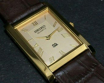 May include: A gold-toned Seiko Quartz watch with a rectangular face and Roman numeral hour markers. The watch has a brown leather strap. The watch face is a light champagne colour with the Seiko logo and a small emblem.