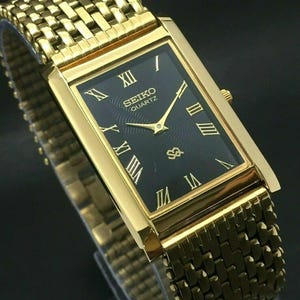May include: A gold-tone rectangular Seiko Quartz watch with a black dial featuring Roman numeral hour markers, gold hands, and a textured centre. The watch includes a matching gold-tone metal link bracelet with a woven, multi-link design pattern.
