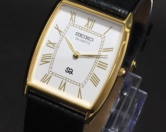 May include: A rectangular Seiko Quartz watch with a gold-tone case and a white face featuring gold Roman numerals. The watch has a black leather strap and a small logo at the bottom of the face.