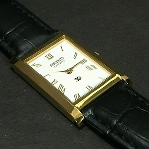 Puede incluir: Un reloj Seiko Quartz rectangular dorado con esfera blanca y correa de cuero negro. La esfera del reloj presenta números romanos y agujas doradas. La correa de cuero negro tiene un patrón texturizado similar al de un cocodrilo.