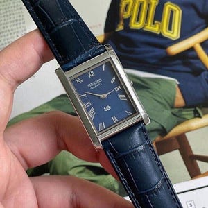 May include: A rectangular wristwatch with a silver-tone case and a dark blue dial. The dial features silver-tone Roman numeral hour markers, silver-tone hands, and the text Seiko Quartz with an SQ logo. It has a dark blue embossed leather strap.