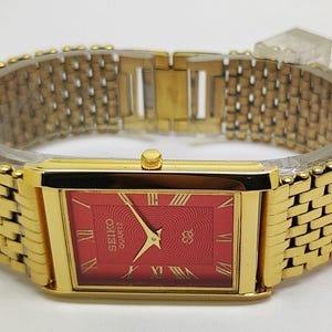 Puede incluir: Un reloj de pulsera rectangular en tono dorado con una esfera roja vibrante con un patrón texturizado, marcadores de hora con números romanos dorados y manecillas doradas. Incluye una pulsera de eslabones de metal dorado a juego.