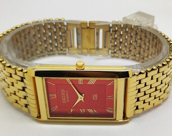 May include: A gold-tone rectangular wristwatch featuring a vibrant red dial with a textured pattern, gold Roman numeral hour markers, and gold hands. The watch includes a matching gold-tone metal link bracelet with a secure clasp mechanism.