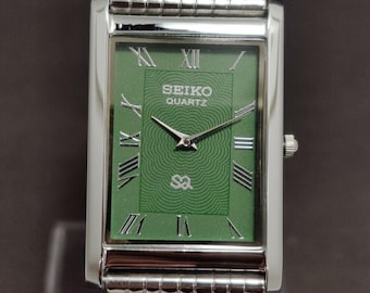 May include: A rectangular Seiko Quartz wristwatch with a green dial featuring a concentric wave pattern, silver Roman numeral hour markers, and silver hands. The watch has a polished silver-tone metal case and a matching multi-link metal bracelet.