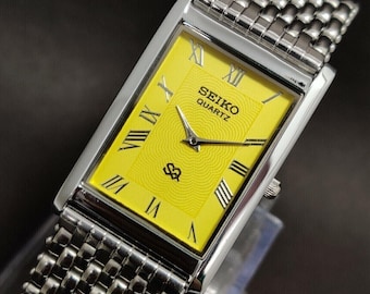 May include: A rectangular Seiko Quartz watch with a bright yellow dial featuring a concentric wave pattern, black Roman numeral hour markers, and silver-tone hands. The watch has a polished silver-tone metal case and a multi-link metal bracelet.