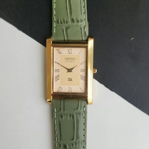 May include: A gold-toned Seiko Quartz watch with a rectangular face and Roman numeral hour markers. The watch has a light-colored face and a green leather band. The band has a textured, crocodile-like pattern.