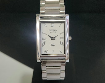 May include: A rectangular silver-tone Seiko Quartz wristwatch with a white dial, black Roman numeral hour markers, and black hands. The watch features a polished metal link bracelet and a crown on the right side of the rectangular case.