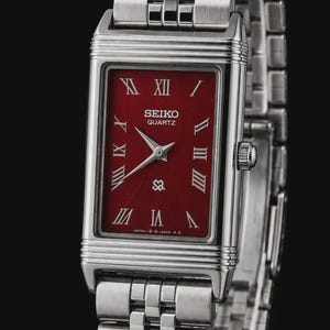 May include: A rectangular Seiko Quartz wristwatch with a deep red dial, silver-toned Roman numeral hour markers, and silver hands. The watch features a polished silver-tone metal case and a multi-link stainless steel bracelet strap.