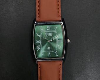 May include: A rectangular Seiko Quartz wristwatch featuring a sunray green dial with silver Roman numeral hour markers, a silver-tone case, and a smooth tan leather strap with visible stitching along the edges of the band.