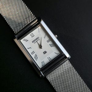 May include: A rectangular Seiko Quartz wristwatch with a silver-tone metal mesh strap. The white dial features black Roman numeral hour markers, a central wavy line pattern, and a small logo above the six o'clock position with two silver hands.