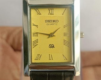 May include: A rectangular Seiko Quartz wristwatch featuring a yellow dial with a concentric wave pattern, gold-tone Roman numeral hour markers, and gold-tone hands. The watch has a polished silver-tone metal case and a black leather strap.