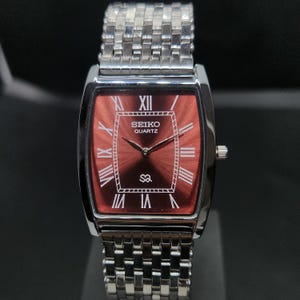 Op de afbeelding: Een rechthoekig Seiko Quartz polshorloge met een dieprode sunray-wijzerplaat en witte Romeinse cijfers. Het horloge heeft een gepolijste zilverkleurige metalen kast en een bijpassende schakelarmband met een geweven textuurontwerp.