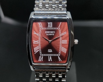 May include: A rectangular Seiko Quartz wristwatch featuring a deep red sunray dial with white Roman numeral hour markers. The watch has a polished silver-tone metal case and a matching multi-link bracelet with a textured, woven-style design.