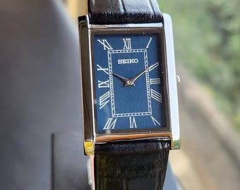 May include: A rectangular Seiko wristwatch featuring a deep blue dial with white Roman numeral hour markers and silver-tone hands. The watch has a polished silver-tone metal case and a black leather strap with a textured crocodile-style pattern.