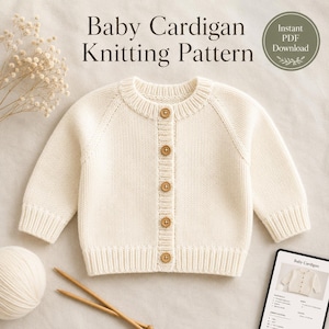 May include: Cream-colored baby cardigan with wooden buttons. The knitting pattern is visible, along with a ball of yarn and knitting needles. The text "Baby Cardigan Knitting Pattern" is displayed at the top of the image.