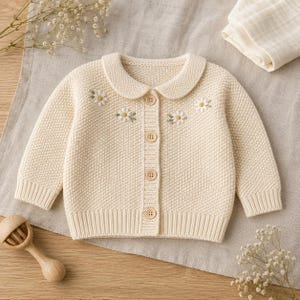 May include: A cream-colored long-sleeve knit cardigan featuring a rounded collar, ribbed cuffs and hem, and four light brown buttons down the center. The upper chest area is decorated with small embroidered white daisy flowers with yellow centers.