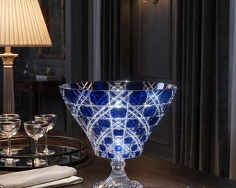 May include: A large, deep, cobalt blue crystal bowl with a wide, flared rim and intricate, intersecting diamond-cut patterns. The bowl sits on a thick, clear, faceted pedestal base, reflecting light through its heavy, ornate glass structure.