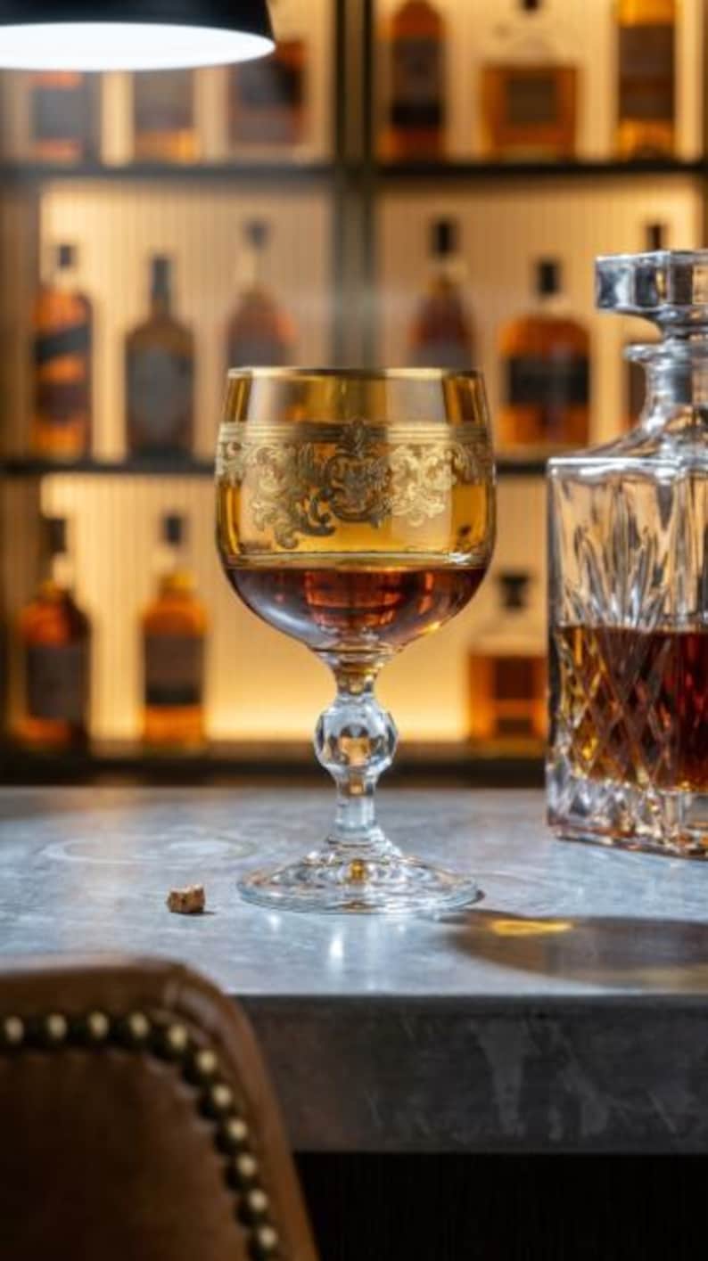 May include: A stemmed glass featuring an amber-tinted bowl with an intricate gold filigree band around the upper portion. The glass is filled with a dark amber liquid and sits on a clear, faceted pedestal base next to a crystal decanter.