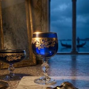 May include: A deep blue glass goblet featuring intricate gold filigree patterns around the rim and a faceted crystal stem, accompanied by a second matching goblet and a pair of vintage brass binoculars resting on a marble slab.