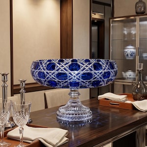 May include: A large, deep, cobalt blue crystal bowl featuring an intricate diamond-cut pattern on its exterior. The bowl is supported by a clear, pedestal-style base with a fluted design, creating a striking contrast between the blue and clear glass.