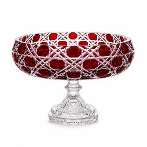 May include: A deep red crystal bowl with a clear glass pedestal base. The bowl features a complex cut-glass pattern of intersecting lines and hexagonal shapes. The clear glass stem and circular base have a vertical ribbed texture and faceted edges.