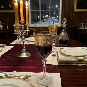 K&ouml;nnte beinhalten: Elegante Tischdekoration mit brennendem Kerzenleuchter, Weingl&auml;sern und einer Karaffe. Das Weinglas hat ein goldenes Filigranmuster. Ein weinroter Tischl&auml;ufer und wei&szlig;e Teller mit Goldrand vervollst&auml;ndigen die Szene. Ein verschneite Stra&szlig;e ist zu sehen.