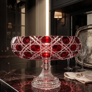 May include: A large, deep red crystal bowl with a wide, circular rim and a complex, diamond-cut geometric pattern. The bowl is supported by a clear, fluted pedestal base with a thick, circular foot featuring intricate, pressed glass detailing.