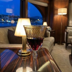 K&ouml;nnte beinhalten: Ein Weinglas mit rotem Wein, verziert mit einem goldenen Muster am oberen Rand. Das Glas steht auf einem dunklen Untersetzer auf einer polierten Holzoberfl&auml;che. Im Hintergrund eine Lampe und eine Aussicht auf den Nachthimmel.