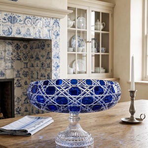 May include: A large, deep cobalt blue glass bowl with a complex, diamond-cut geometric pattern on the exterior. The bowl is supported by a clear, fluted glass pedestal base that features a circular, scalloped edge at the very bottom.