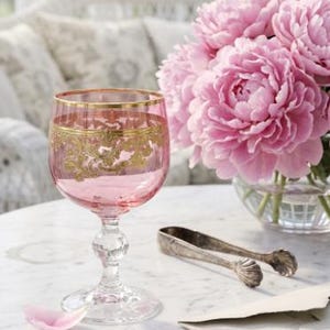 May include: A pink glass goblet featuring a gold-colored decorative band around the rim and a clear glass stem with a spherical accent. The goblet is positioned next to a bouquet of pink peonies, silver-toned metal tongs, and a light-colored envelope.