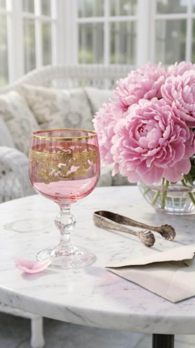 May include: A pink glass goblet featuring a gold-colored decorative band around the rim and a clear glass stem with a spherical accent. The goblet is positioned next to a bouquet of pink peonies, silver-toned metal tongs, and a light-colored envelope.