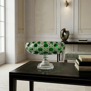 May include: A green and clear crystal bowl with a pedestal base. The bowl has a diamond-cut pattern with green accents. The bowl is sitting on a black table in a well-lit room. The bowl is a decorative item.