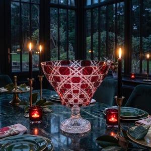 May include: A large, deep, ruby red cut glass bowl with a clear crystal pedestal base. The bowl features an intricate, geometric cross-hatch pattern etched into the glass, creating a faceted appearance that catches the light from nearby candles.