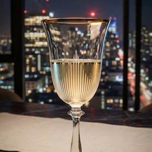 May include: A clear champagne glass with a gold rim, filled with a light-coloured liquid. The glass has vertical etched lines and sits on a long stem. The background shows a city skyline at night with a chandelier.