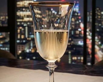 May include: A clear champagne glass with a gold rim, filled with a light-colored liquid. The glass has vertical etched lines and sits on a long stem. The background shows a city skyline at night with a chandelier.