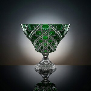 May include: A green crystal bowl with a pedestal base. The bowl has a geometric pattern and is reflected in the surface below. The bowl is a decorative piece.