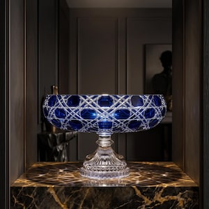 May include: A deep blue glass bowl with a wide, circular rim and a complex, intersecting diamond-cut pattern. The bowl is supported by a clear, fluted pedestal base with a scalloped edge, reflecting light through its intricate, faceted surface.