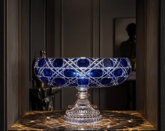 May include: A deep blue glass bowl with a wide, circular rim and a complex, intersecting diamond-cut pattern. The bowl is supported by a clear, fluted pedestal base with a scalloped edge, reflecting light through its intricate, faceted surface.