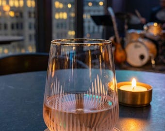 May include: A stemless wine glass with a gold rim and etched vertical lines, filled with a light-colored liquid, sits on a leather coaster. A lit candle in a gold holder is next to the glass. The background shows a city skyline and a neon sign.