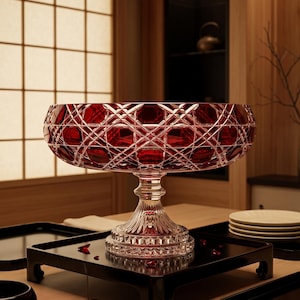 May include: A deep red cut glass pedestal bowl featuring a complex geometric cross-hatch pattern on the exterior. The bowl sits on a clear, fluted glass base with a scalloped edge, showcasing a vibrant ruby hue and reflective, faceted surfaces.