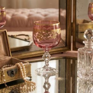 May include: A pink glass goblet featuring a gold-colored ornate leaf pattern around the rim and a clear, faceted stem. The glass is positioned on a reflective surface alongside a small brass-toned box with pearls and a clear crystal decanter.