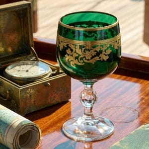 May include: A green glass goblet featuring an intricate gold filigree pattern around the rim and bowl, supported by a clear, faceted crystal stem and a circular base, standing upright on a polished wooden surface next to a brass nautical instrument.