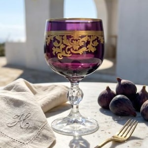 May include: A deep purple glass goblet featuring an intricate gold filigree band around the upper rim. The stem is clear with a faceted, diamond-like cut design, and the base is a wide, clear circular disc. The glass sits on a marble surface.