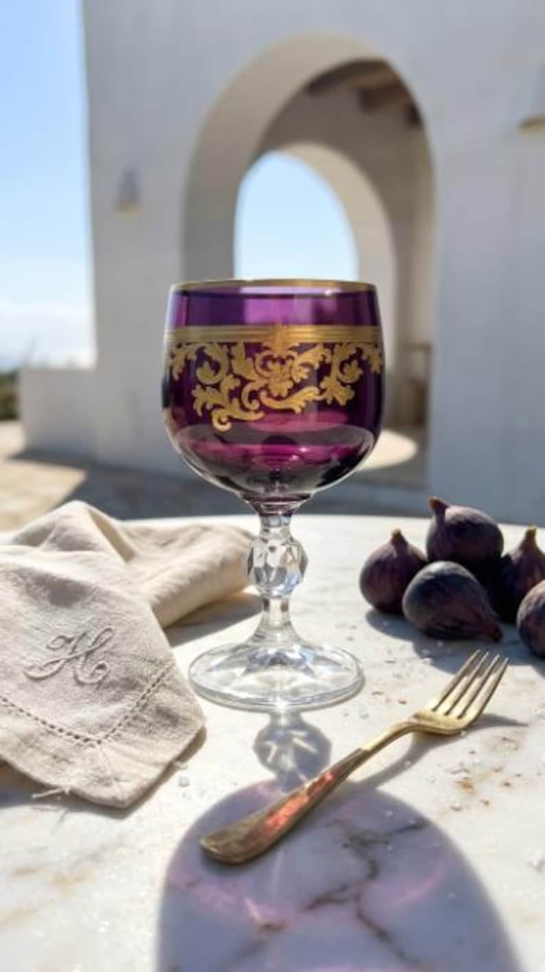 May include: A deep purple glass goblet featuring an intricate gold filigree band around the upper rim. The stem is clear with a faceted, diamond-like cut design, and the base is a wide, clear circular disc. The glass sits on a marble surface.