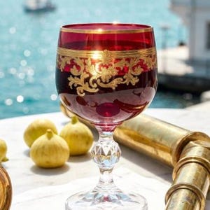 May include: A deep red glass goblet featuring intricate gold filigree patterns around the rim, supported by a clear, faceted crystal stem and base. The glass is positioned alongside a brass telescope and a gold-toned pocket watch.