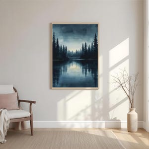 May include: A framed landscape art print depicts a serene lake scene with dark evergreen trees lining the water's edge. The artwork features a muted color palette of blues and greens, creating a tranquil atmosphere. The print is displayed on a neutral-colored wall.