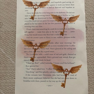 May include: Open book page with text from Chapter Fourteen. Three golden keys with wings are superimposed on the page. The text includes the words "Harry", "Ron", and "Quidditch". The page is slightly stained with a pink hue.