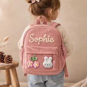 Peut inclure: Sac à dos en velours côtelé rose avec le nom "Sophie" brodé sur le devant. Le sac à dos comporte une poche avant, une poche latérale et des appliqués décoratifs de fleurs et de lapin. Bretelles réglables.