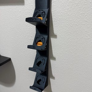 Puede incluir: Un estante de almacenamiento negro montado en la pared con cuatro estantes, cada uno diseñado para sostener una consola de juegos. Los estantes tienen un diseño circular con forma de 'X' en el centro. El estante está montado en una pared blanca.